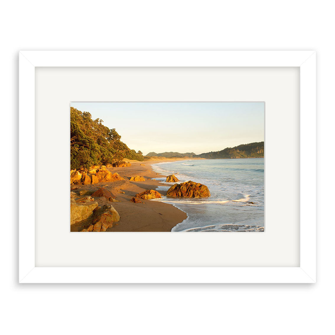 Hot Water Beach - RP122 is a framed Reuben Price print featuring a sandy shore with rocks, gentle waves, and trees under a clear sky. Displayed in a white mat and frame on archival quality matte paper.