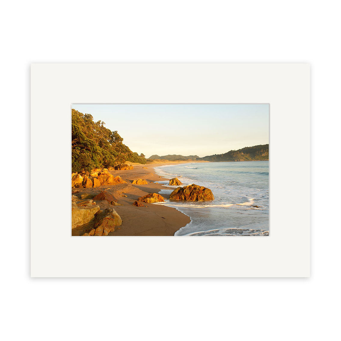 Hot Water Beach - RP122, part of the Reuben Price prints collection, shows a sandy beach with rocks and waves, bordered by trees and hills. Presented in a white mat frame, it is printed on archival quality matte paper for lasting beauty.