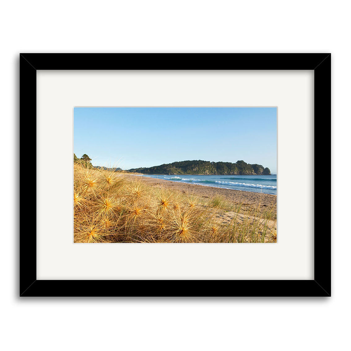 Hot Water Beach - RP123 is a framed photographic print by Reuben Price, featuring dried grass on sand, gentle waves, and a tree-covered headland beneath a clear blue sky, printed on archival quality matte paper.