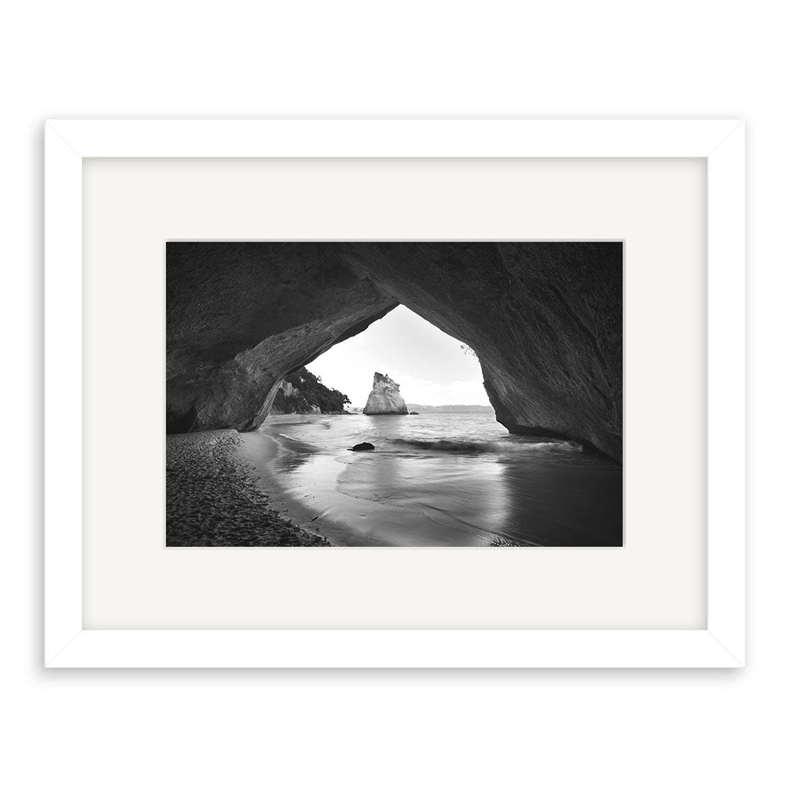 Cathedral Cove - RP125: Black-and-white photo of a natural rock arch in New Zealand, showcasing beach and ocean views with a large rock. Presented as a photographic print in a white frame with archival quality matte paper mat.