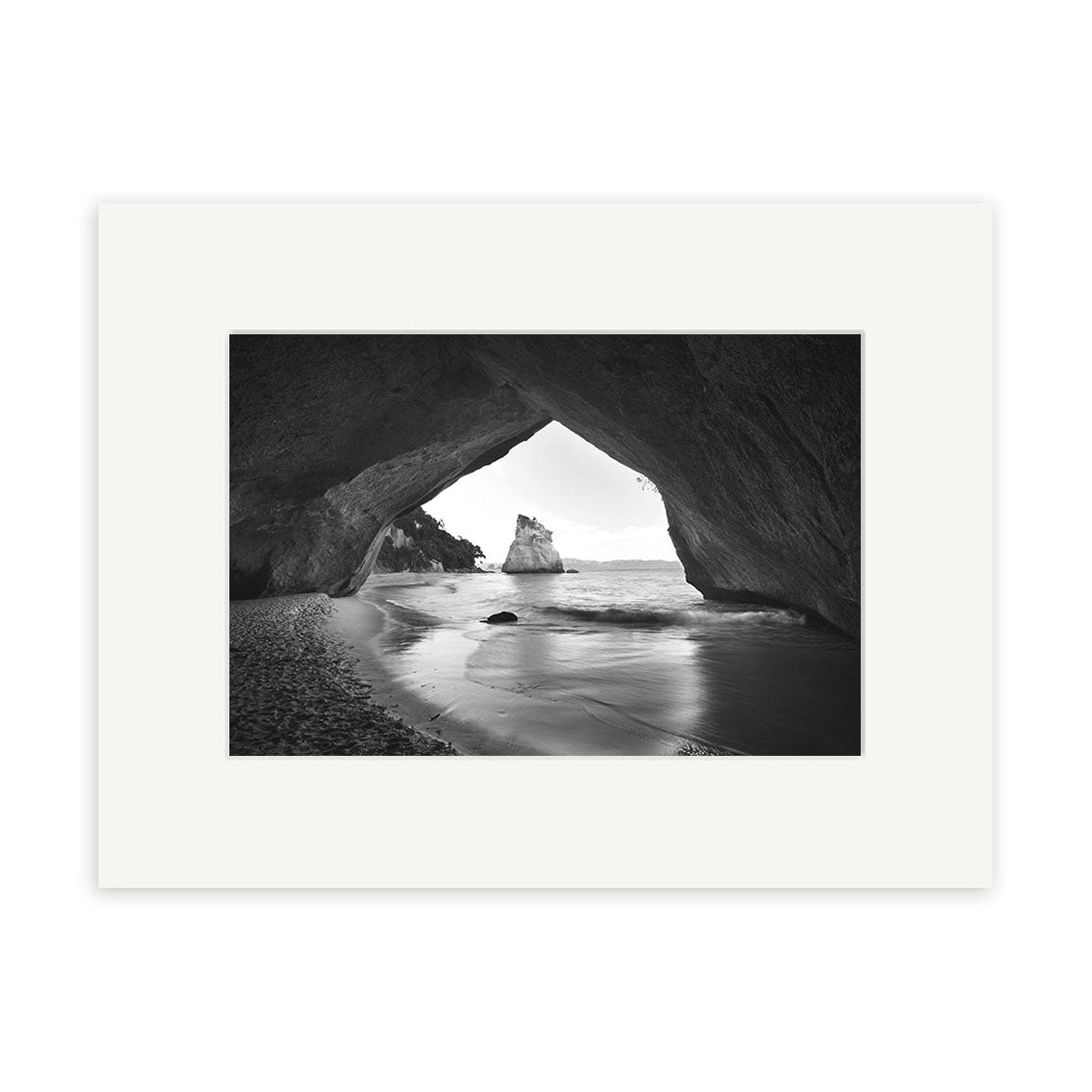 Cathedral Cove - RP125: Black and white photo of a coastal rocky cave opening onto a sandy New Zealand beach, with a large sea rock visible through the entrance. Available as archival quality matte paper photographic prints.