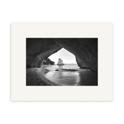 Cathedral Cove - RP125: Black and white photo of a coastal rocky cave opening onto a sandy New Zealand beach, with a large sea rock visible through the entrance. Available as archival quality matte paper photographic prints.