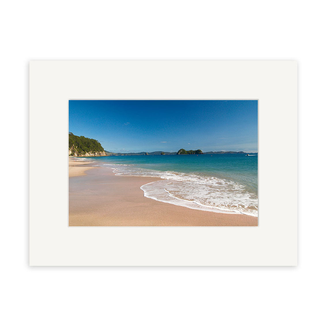 Hahei Beach - RP126 features a sandy shore with gentle waves, clear blue water, and a rocky coastline beneath a bright sky. The photo is printed on archival matte paper and framed with a wide white mat for lasting beauty.