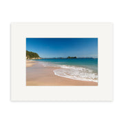 Hahei Beach - RP126 features a sandy shore with gentle waves, clear blue water, and a rocky coastline beneath a bright sky. The photo is printed on archival matte paper and framed with a wide white mat for lasting beauty.