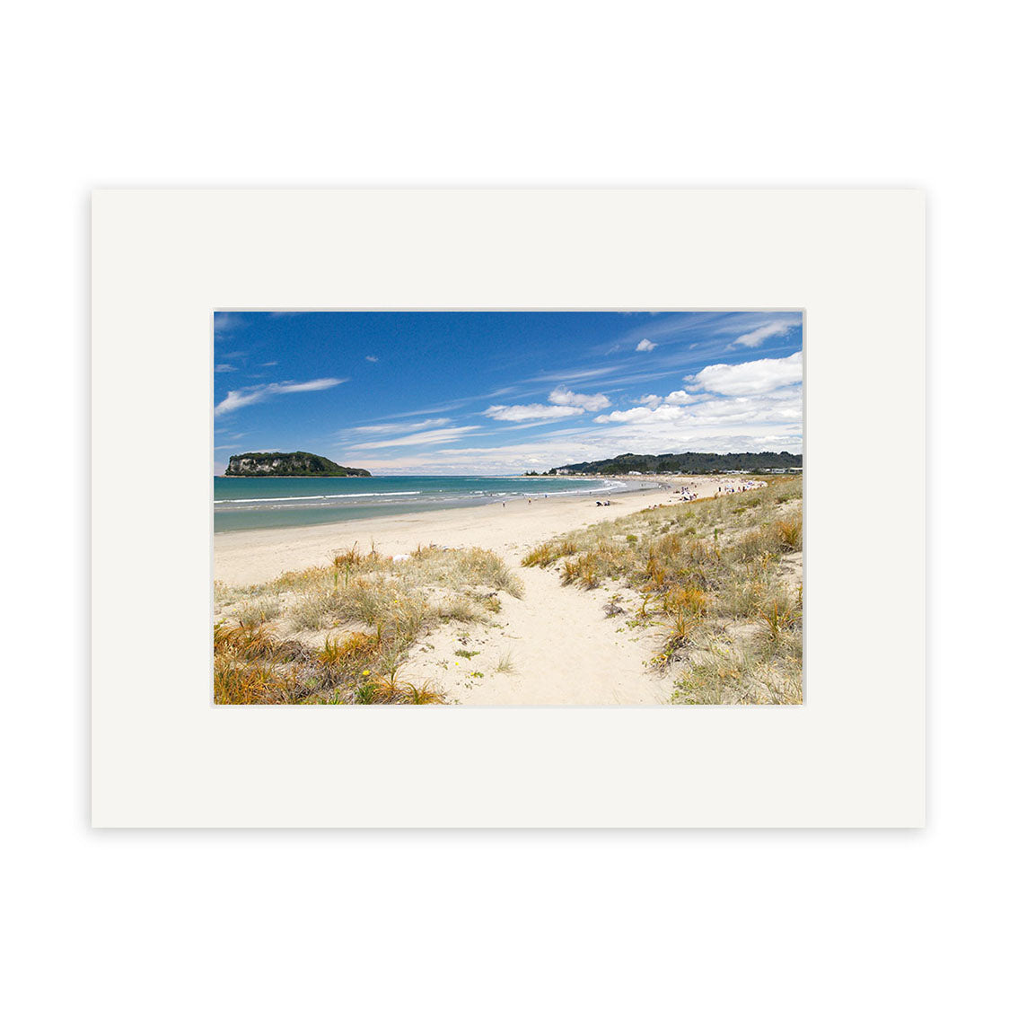 The Whangamata - RP128 print features a sandy beach with grassy dunes, calm blue water, and a distant peninsula beneath scattered clouds. Presented with a white matte, its perfect for showcasing New Zealand’s natural beauty.
