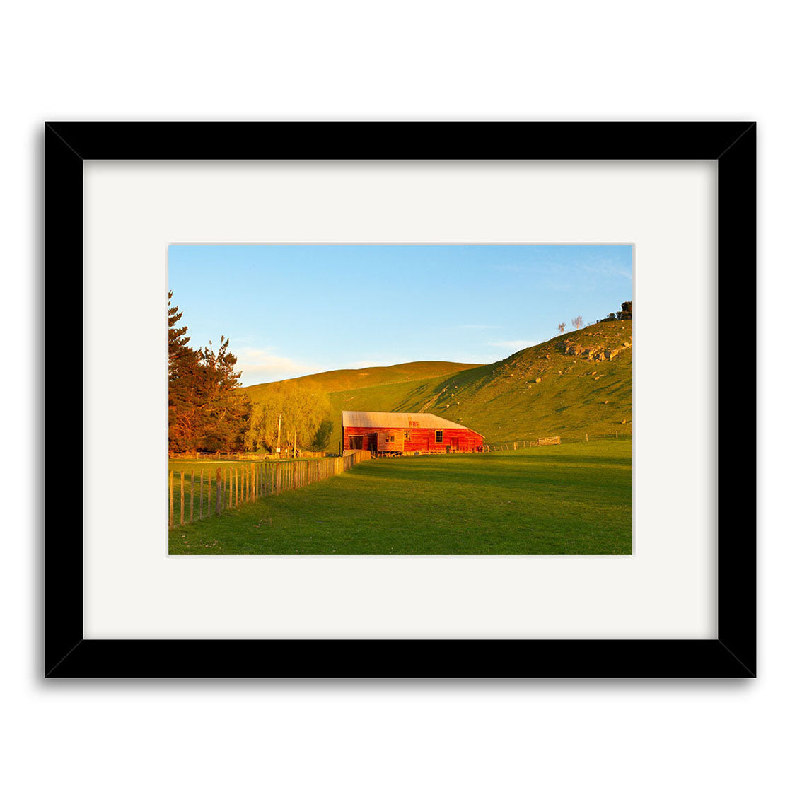 The Havelock North Red Barn - RP133 is a framed photo by Reuben Price showing a red barn in a sunlit green New Zealand field with hills, trees, and a fence—ideal for fans of scenic photographic prints.