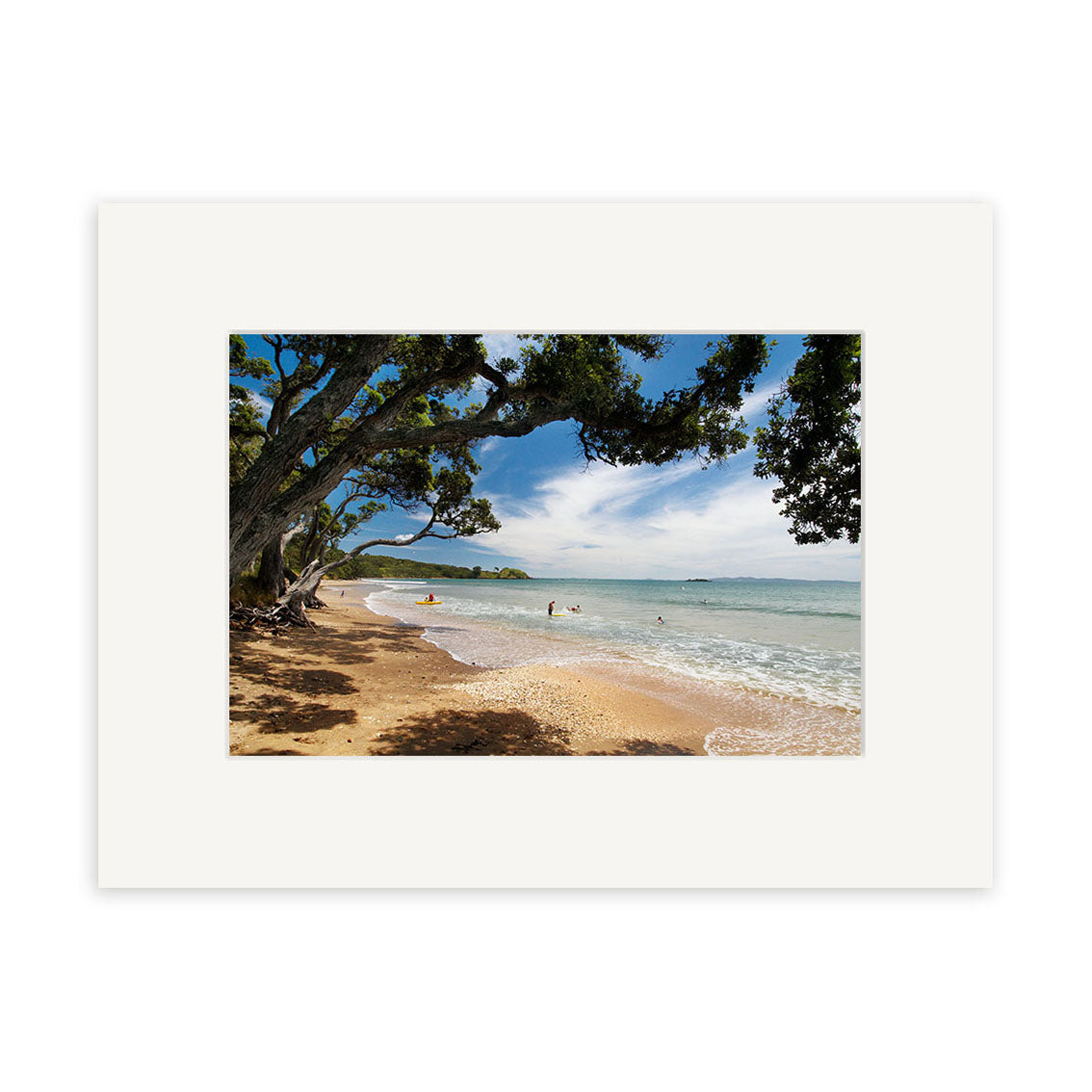 The scene from Coopers Beach - RP137 shows gentle waves, swimmers, and leafy trees along a sandy New Zealand shore under a partly cloudy sky. Beautifully framed with a white mat, its ideal for archival quality matte paper photographic prints.