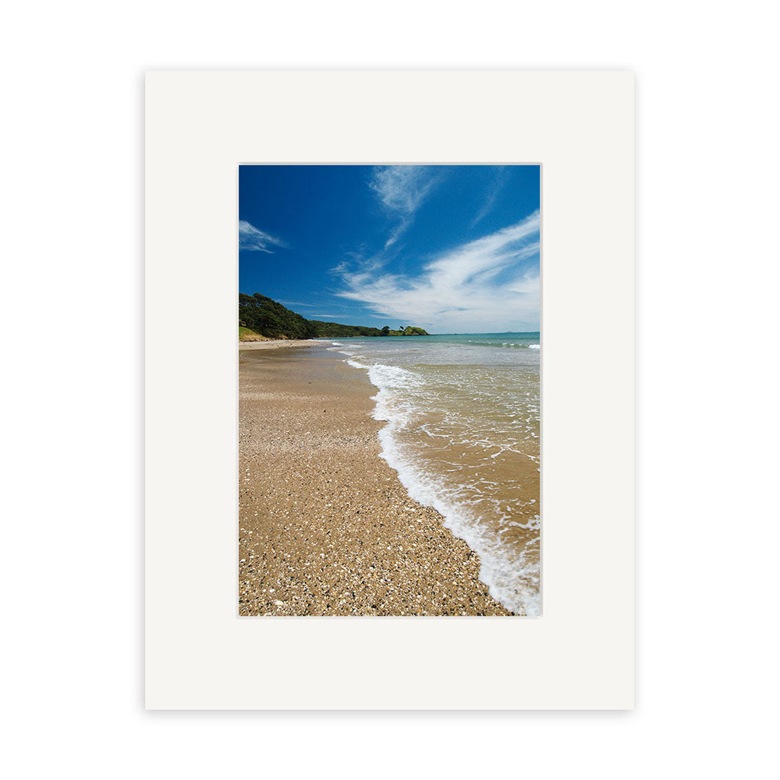 A sandy beach with gentle waves and green trees under a blue sky features in Coopers Beach - RP138, a photographic print by Reuben Price of New Zealand’s natural beauty, centered within a plain white mat frame.