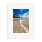 A sandy beach with gentle waves and green trees under a blue sky features in Coopers Beach - RP138, a photographic print by Reuben Price of New Zealand’s natural beauty, centered within a plain white mat frame.