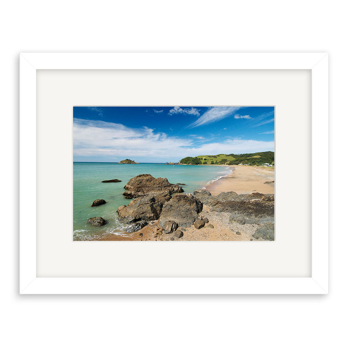 Te Ngaire - RP140 is a framed photograph of a New Zealand coastal landscape featuring sandy beach, scattered rocks, turquoise water, grassy hills, and a blue sky. Printed on archival matte paper with a white frame and wide mat border.