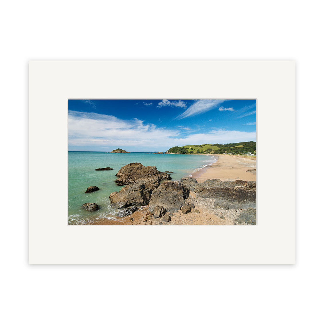 Te Ngaire - RP140: A photographic print of a New Zealand sandy beach with rocks, turquoise water, green hills, and blue sky. Presented with a wide white mat on archival quality matte paper.