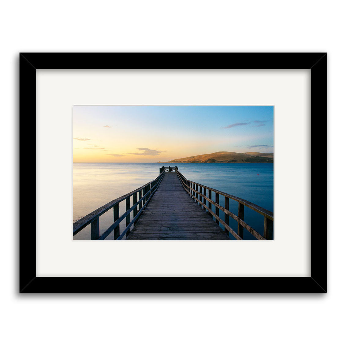 Omapere Wharf - RP141 is a framed photographic print featuring a wooden pier reaching over calm New Zealand waters at sunset. The black frame and white mat accent the image, printed on archival-quality matte paper for enduring beauty.