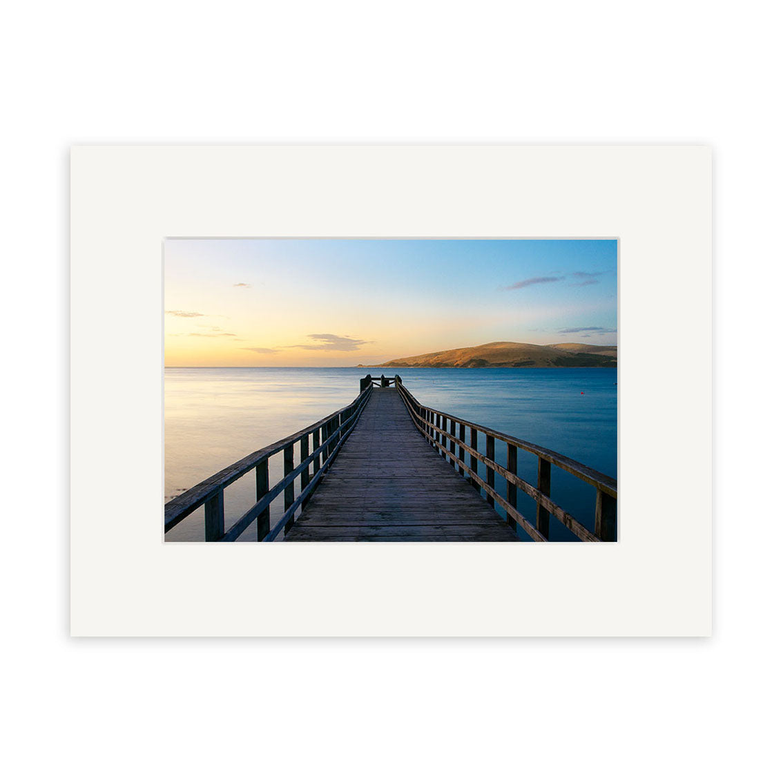 Omapere Wharf - RP141 captures a wooden pier stretching over tranquil water toward distant hills at sunset in New Zealand. Beautifully framed with a white mat, its ideal for photographic prints on archival quality matte paper.
