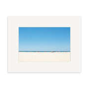 The Pakiri Beach - RP142 framed photographic print by Reuben Price showcases a sweeping view of white sand, distant figures, umbrellas, and clear blue sky—bringing the serene beauty of New Zealand’s coastline to your home.