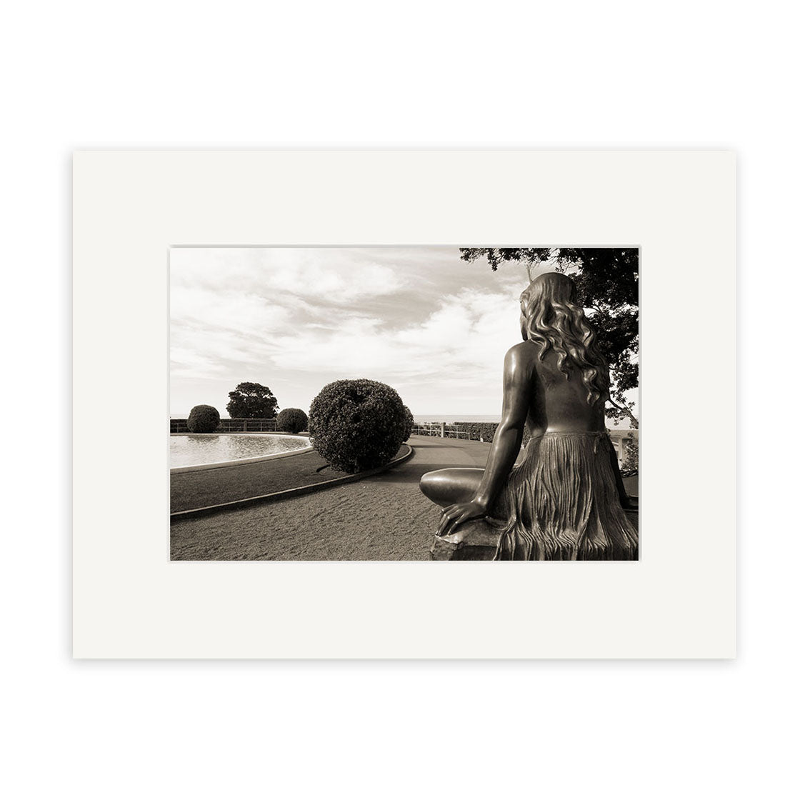 A black-and-white print of Pania of the Reef - RP144 shows a long-haired seated woman statue overlooking a formal New Zealand garden with trimmed bushes, path, and pond beneath partly cloudy skies; framed with a white mat.