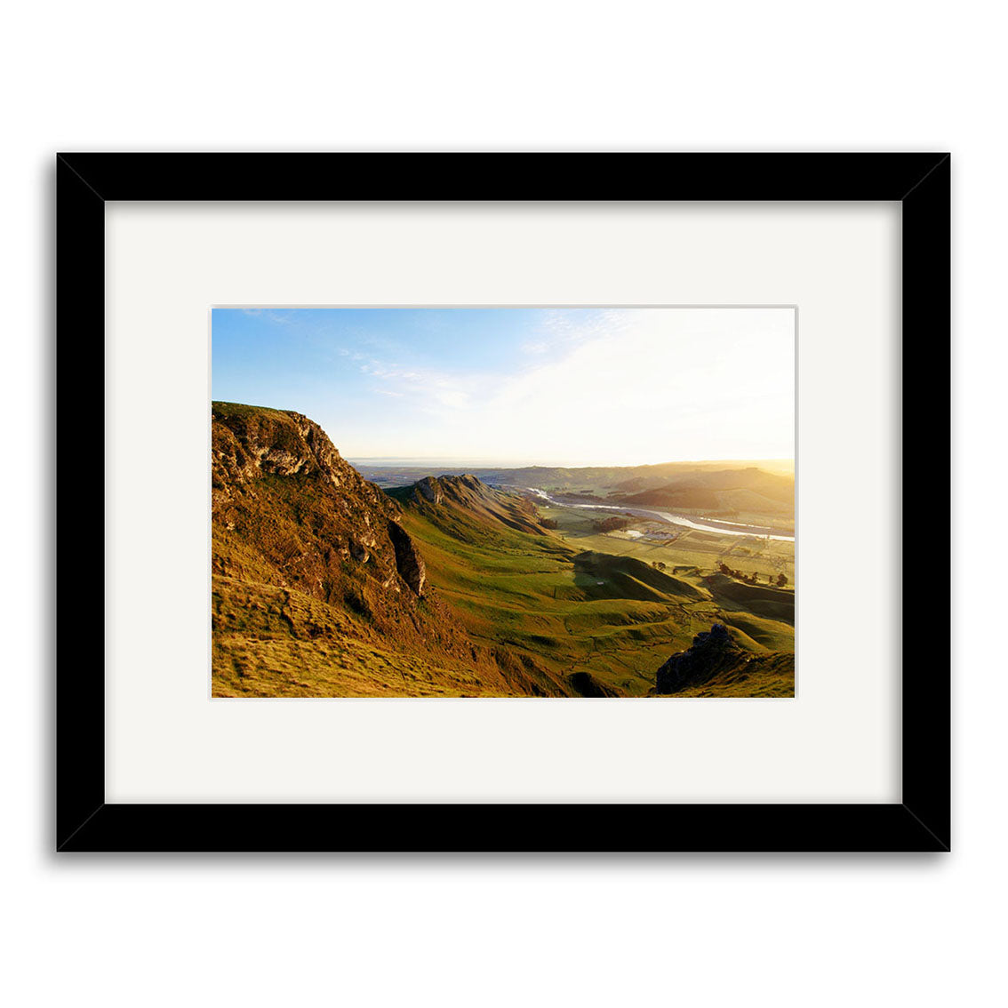 Te Mata Sunrise - RP145 is a framed landscape photograph of New Zealand’s green hills, rocky cliffs, and a sunlit valley. The black frame and white mat enhance the archival quality of this stunning photographic print.