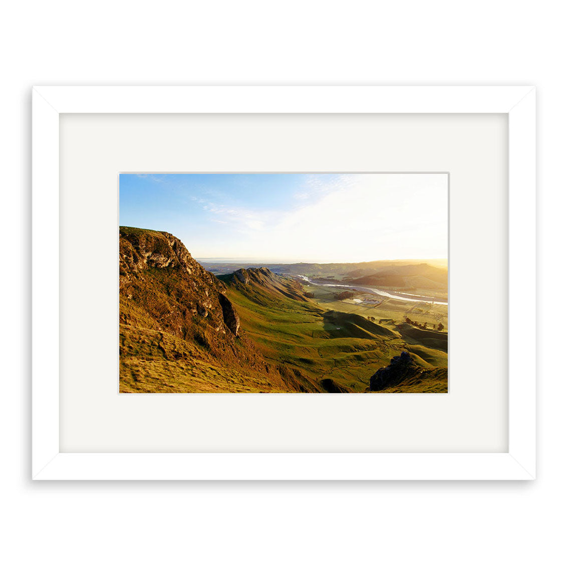 Te Mata Sunrise - RP145: A framed archival-quality photo print of a sunlit New Zealand landscape with grassy hills, rocky cliffs, and a distant valley under a clear sky. Features a white frame with wide mat.