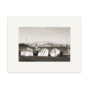 Little Shoal Bay - RP149: Black-and-white photo of upside-down boats on grass in New Zealand, with a bridge and tall tower in the background under a partly cloudy sky. Available as archival quality matte paper print.
