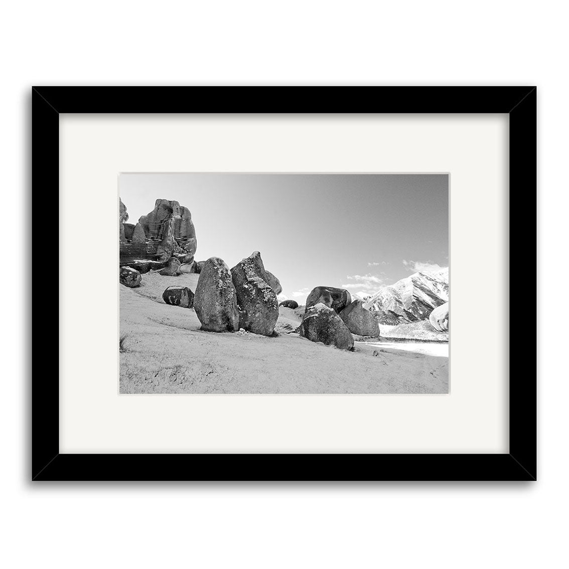 Castle Hill Station - RP151: A framed black-and-white photo of rock formations on a sloped New Zealand landscape, with snow-capped mountains and a partly cloudy sky, expertly printed on archival matte paper.