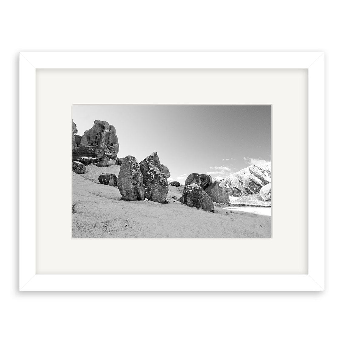 Black and white photo Castle Hill Station - RP151 features large rocky boulders on a grassy slope with snow-capped New Zealand mountains, displayed in a white frame with wide mat and printed on archival quality matte paper.