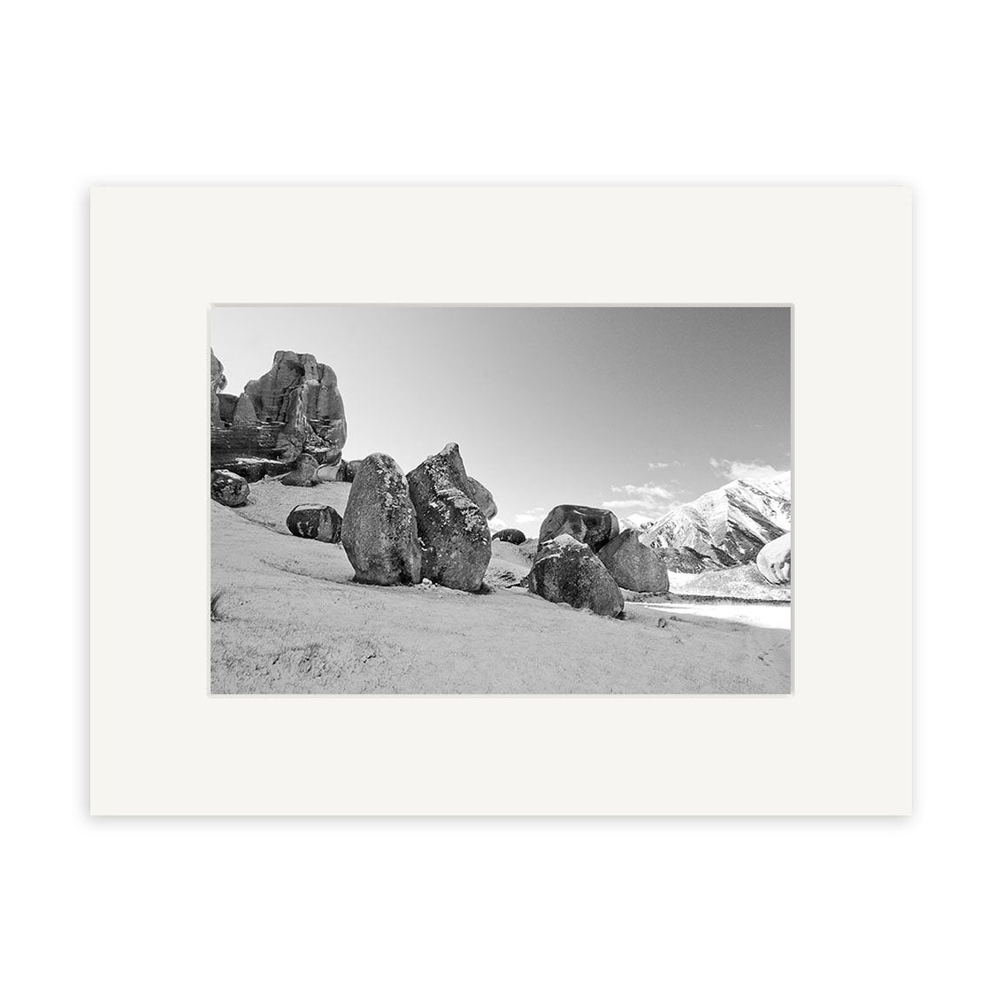 “Castle Hill Station - RP151” is a black and white photographic print of large rock formations on a sandy slope with snowy mountains behind, presented in a white mat and frame. Printed on archival matte paper, it captures New Zealand’s striking scenery.