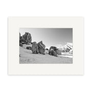 “Castle Hill Station - RP151” is a black and white photographic print of large rock formations on a sandy slope with snowy mountains behind, presented in a white mat and frame. Printed on archival matte paper, it captures New Zealand’s striking scenery.