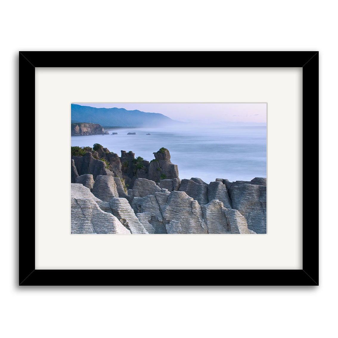 The Punakaiki Sea and Rocks - RP152 is a framed photographic print by Reuben Price, showcasing layered rock formations on New Zealand’s coast with calm water and hills under a blue sky. Features a black frame and white mat.