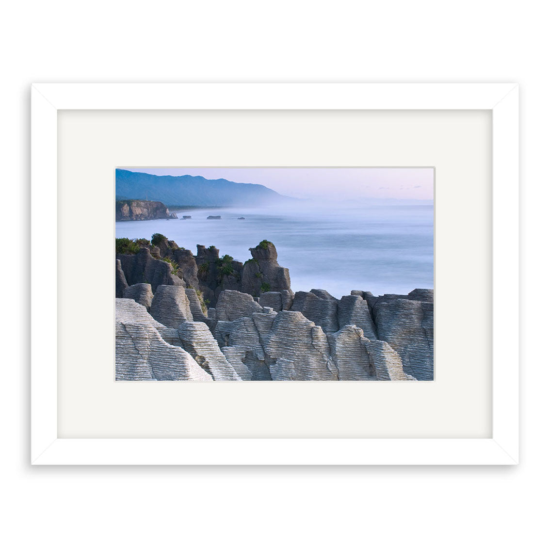 The Punakaiki Sea and Rocks - RP152 framed photographic print by Reuben Price features layered New Zealand rock formations, misty water, and distant mountains beneath a blue sky, all seen from an elevated viewpoint.
