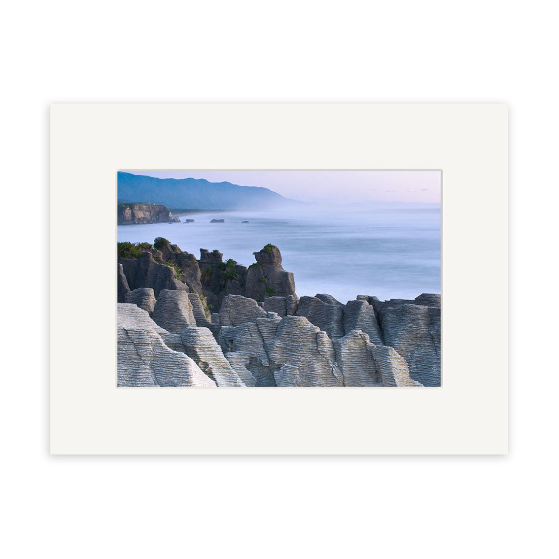 Punakaiki Sea and Rocks - RP152: Striking framed photo by Reuben Price showcasing layered coastal rock formations, ocean mist, distant mountains, and New Zealand’s rugged coastline.