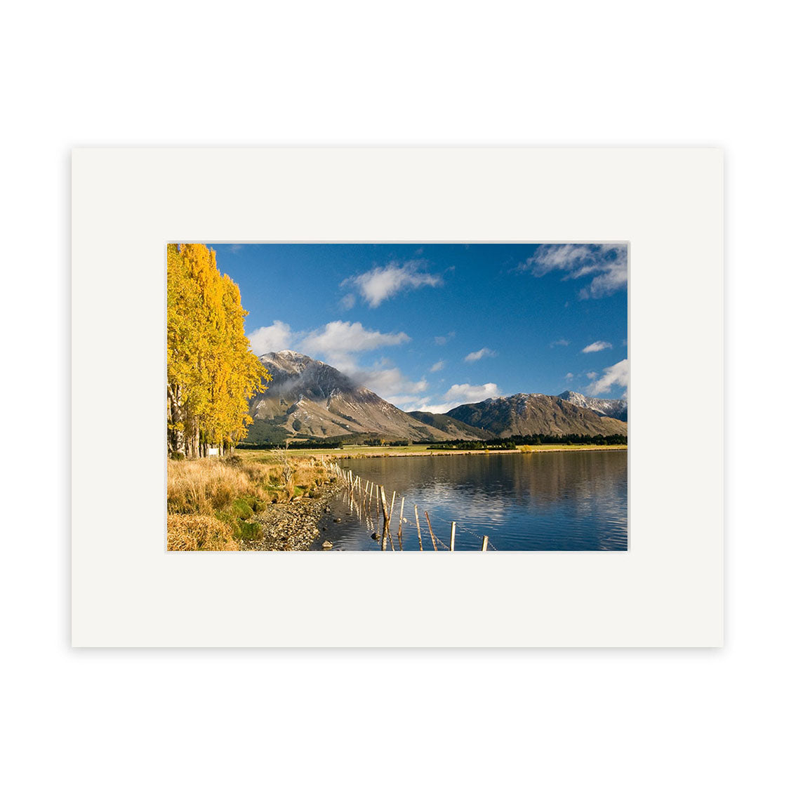 “Lake Grasmere – RP153” is an archival quality photographic print showcasing a New Zealand landscape with a lakeside, yellow-leaved trees, distant mountains, blue sky with clouds, and a wooden fence extending into the water.