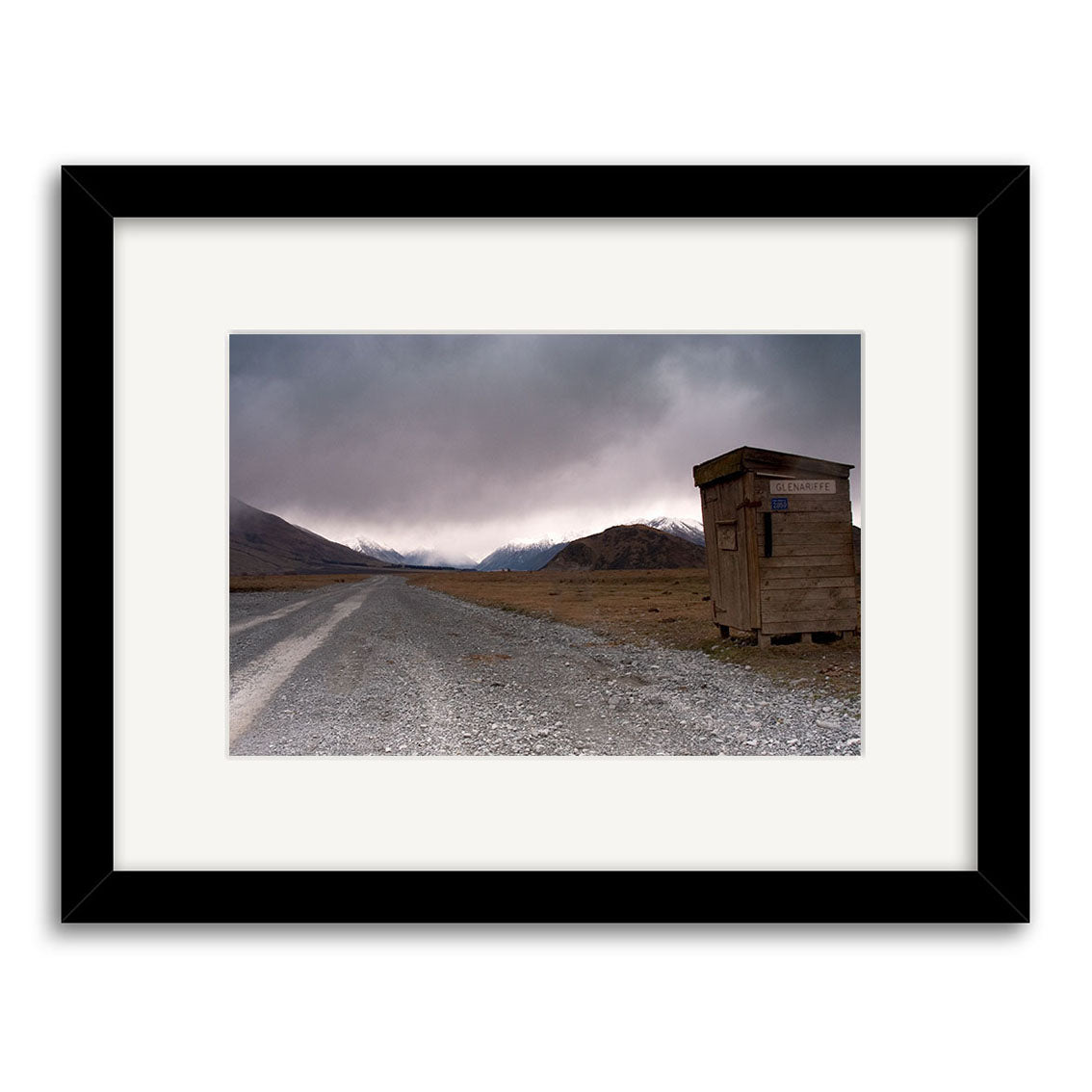 A gravel road winds through a barren, mountainous New Zealand landscape beneath cloudy skies in Glenariffe - RP155, featuring a wooden Immigration Checkpoint shed—ideal for archival quality matte paper photographic prints.