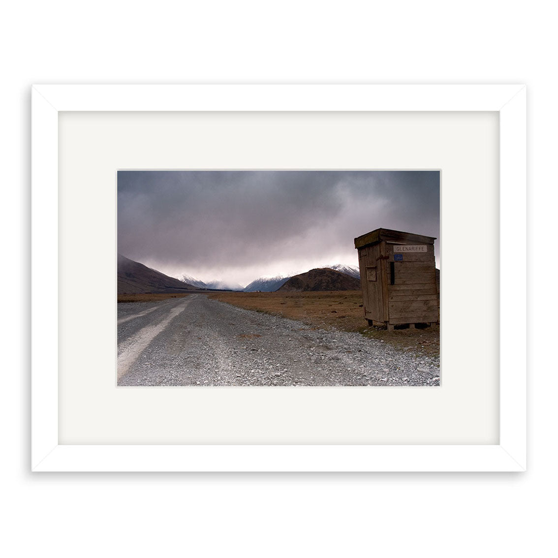 A gravel road leads into a barren, mountainous New Zealand landscape beneath cloudy skies. To the right stands a small wooden structure with a sign, highlighting the remote and desolate scene captured in Glenariffe - RP155.
