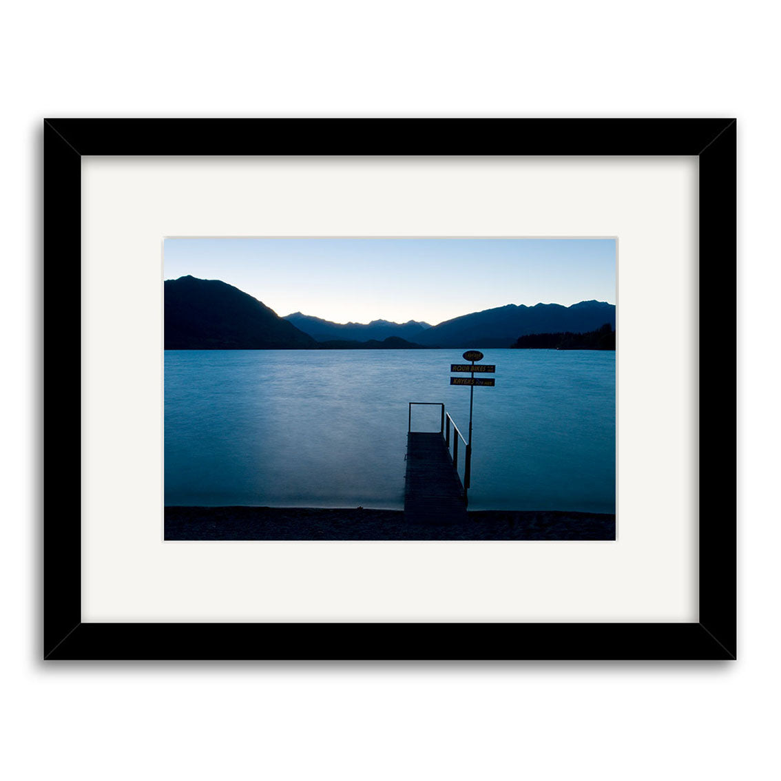 The Lake Wanaka Jetty - RP158 is a framed photographic print featuring a tranquil, dimly lit New Zealand lake at dusk, mountains in the distance, and a small dock with a signpost and railing extending into the water.