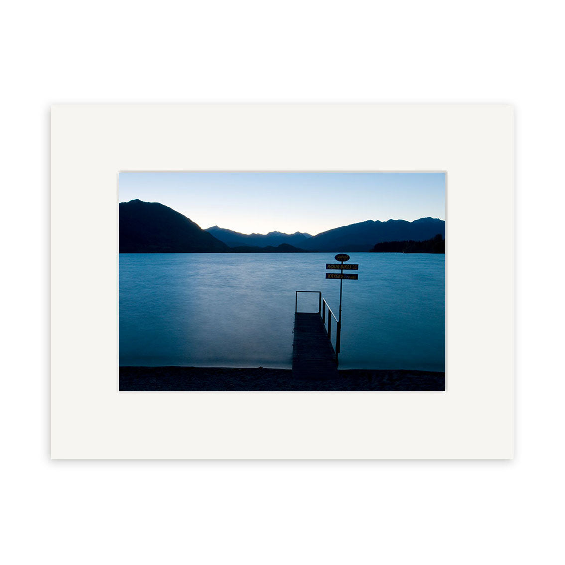 Lake Wanaka Jetty - RP158 captures a tranquil New Zealand lake at dusk, with a dock and silhouetted mountains. A sign marks the docks end. This serene image is ideal for photographic prints on archival quality paper.