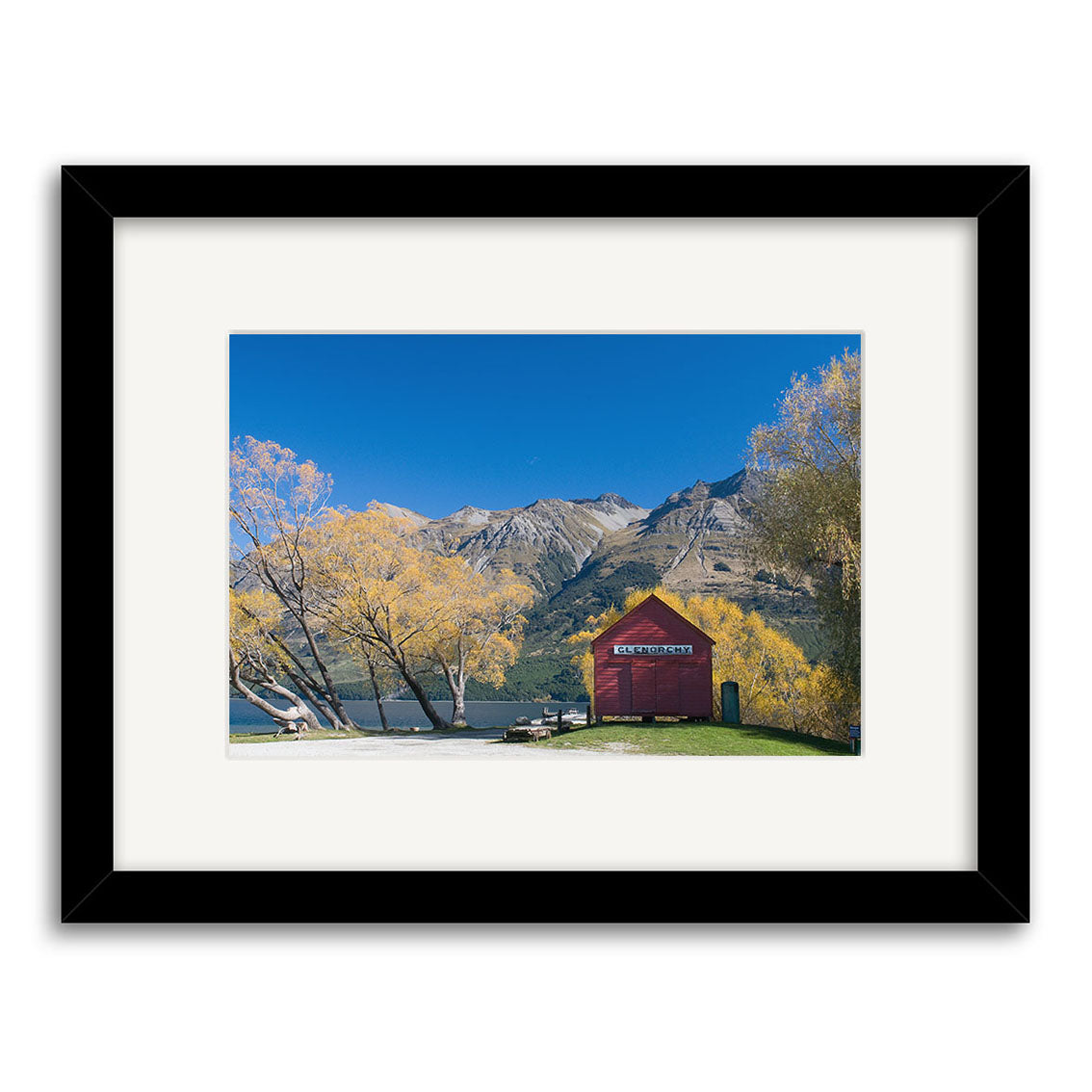 Glenorchy in Autumn - RP159 captures a red shed by a lake, yellow trees, and mountains under a blue sky. This New Zealand scene is presented with a white mat, black frame, and printed on archival matte paper for lasting quality.