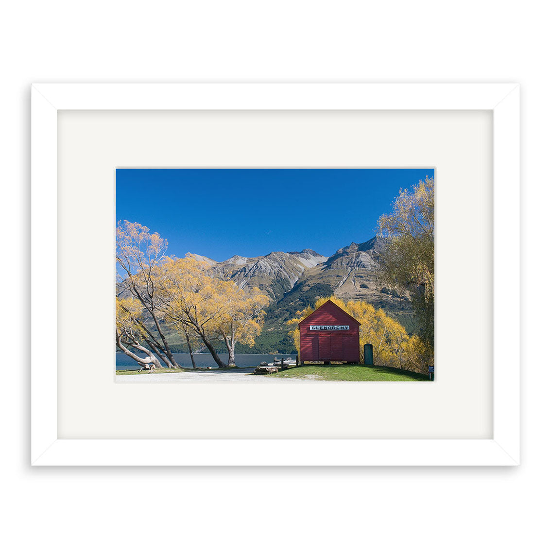 The Glenorchy in Autumn - RP159 framed photographic print shows a red shed by a New Zealand lake, yellow-leaved trees, and mountains under a blue sky, reproduced on archival quality matte paper for lasting beauty.