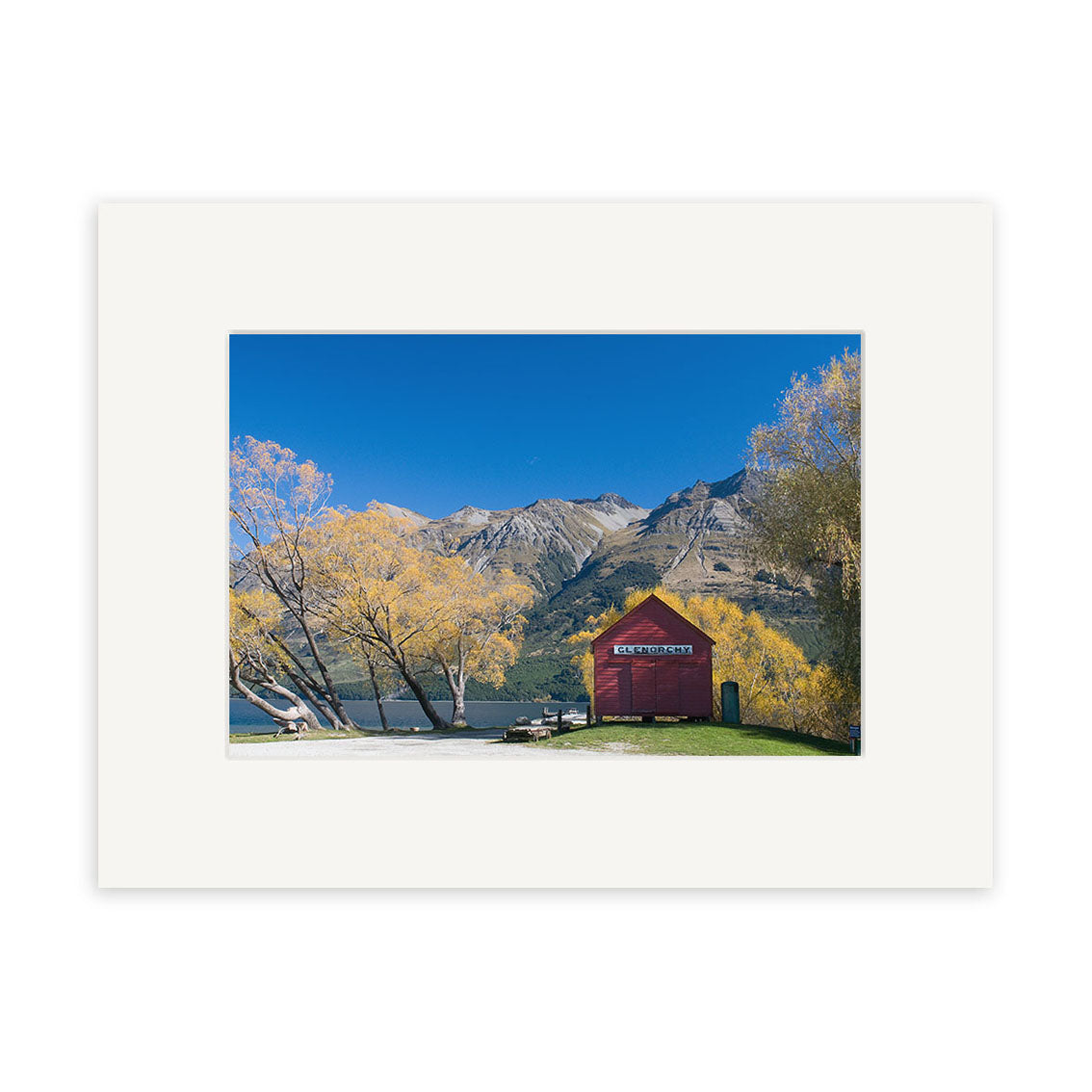 The photographic print Glenorchy in Autumn - RP159 features a red boathouse by a lake with yellow trees and mountains beneath a blue sky, bordered by a white mat on archival-quality matte paper.
