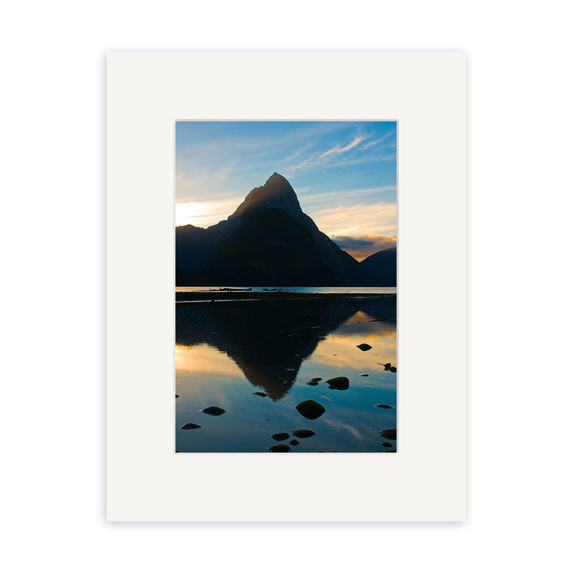 Mitre Peak - RP160 features a mountain peak silhouetted against a sunset sky, reflected in calm water with scattered rocks. This New Zealand scene is presented on archival quality matte paper and framed with a white mat.