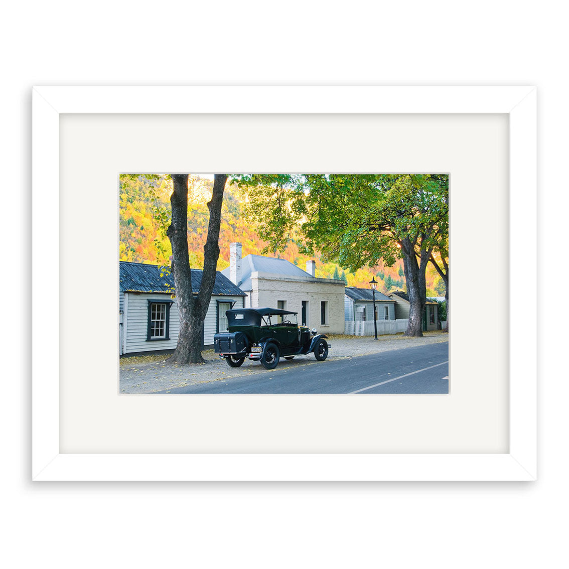 Arrowtown in Autumn - RP162: A vintage black car on a quiet, tree-lined street with historic buildings under a clear sky, beautifully framed and printed on archival matte paper—ideal for lovers of timeless photographic prints.