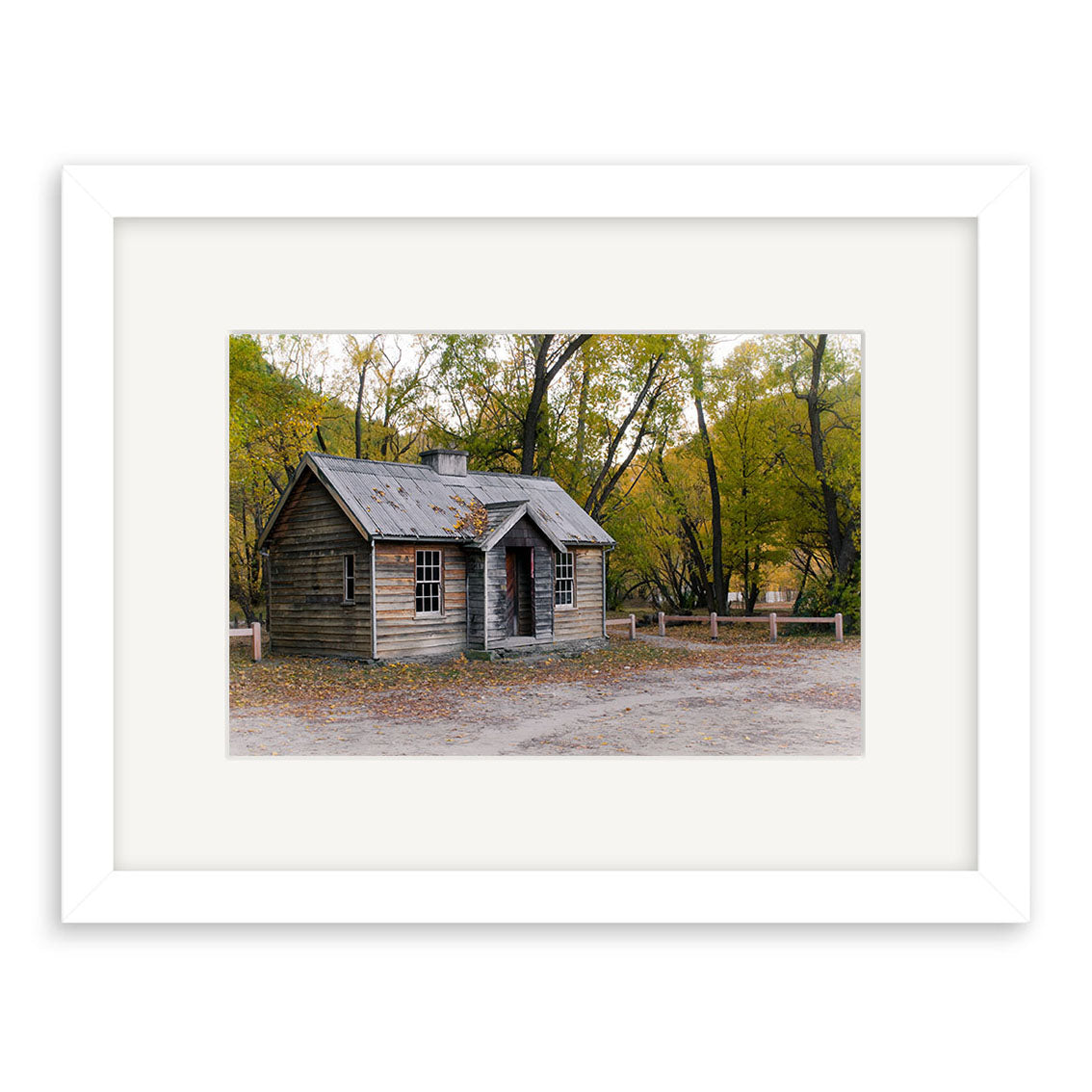 The Arrowtown Old House - RP163 is a framed photographic print on archival paper, showing a weathered wooden house with a tin roof, surrounded by lush New Zealand trees, with a front porch and multiple windows over dirt ground.