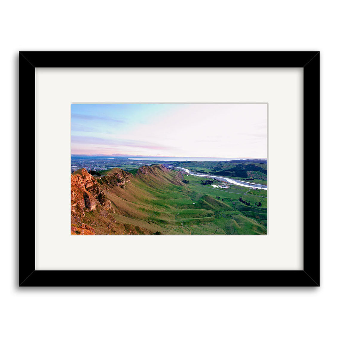Te Mata - RP165 is a stunning photographic print featuring green hills, rocky ridges, and a river or lake beneath a partly cloudy New Zealand sky. The archival-quality print comes framed in black with a white mat for an elegant finish.