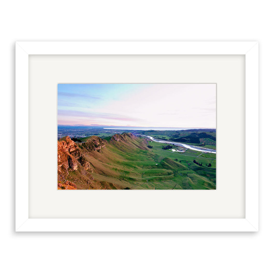 “Te Mata - RP165” is a framed archival quality photographic print of green, hilly New Zealand landscapes with rocky ridges and open fields beneath a partly cloudy sky, displayed with a white mat and simple white frame.