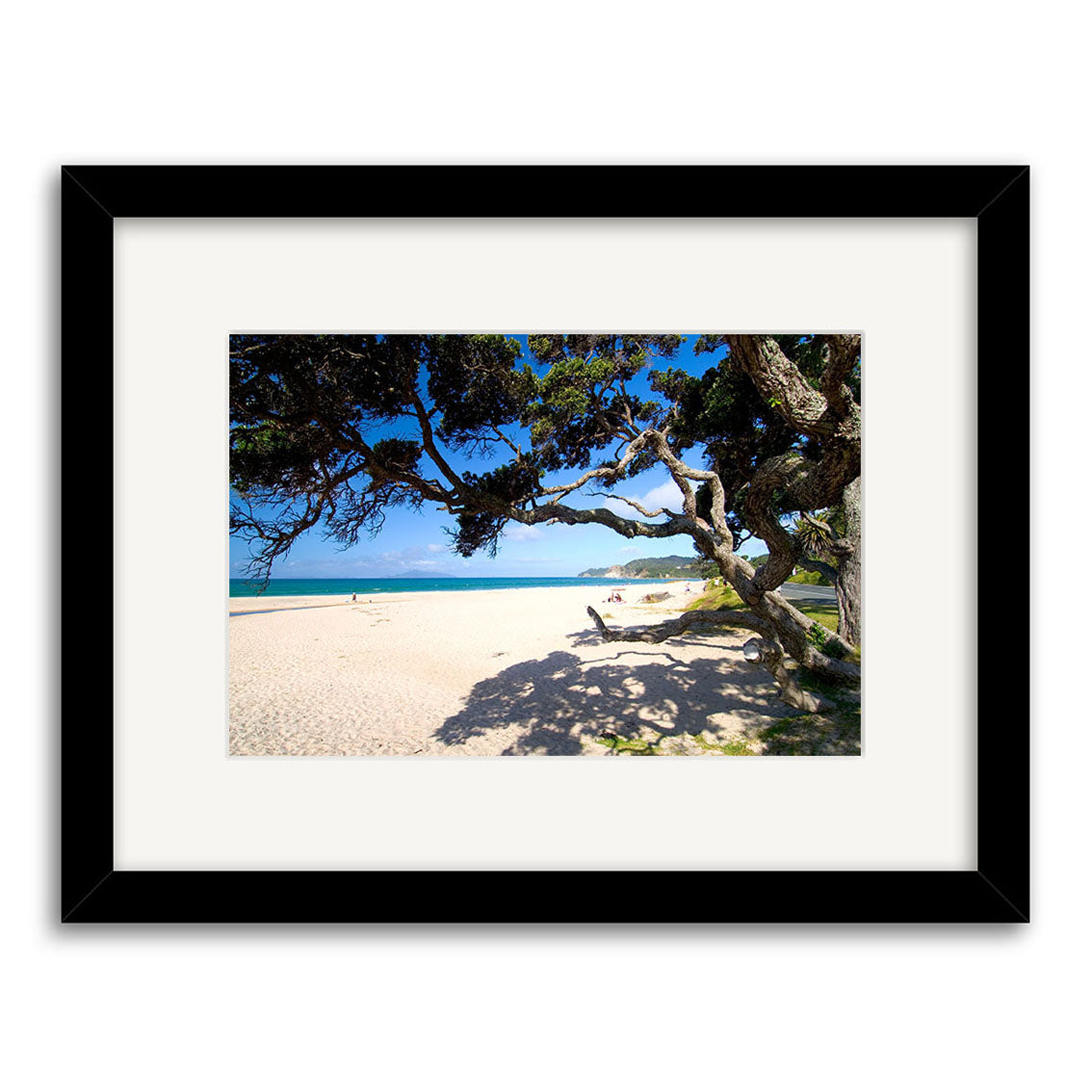 Langs Beach - RP168 is a framed photographic print on archival paper, featuring a sandy beach under blue skies and ocean with a shady tree, centered in a wide white mat and black frame—showcasing New Zealand’s beauty.