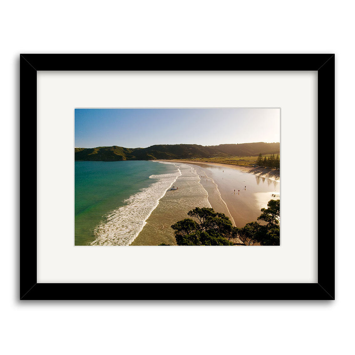 Matauri Bay - RP169: A framed Reuben Price print featuring gentle waves, sandy shoreline, distant hills, and walkers under a clear sky—New Zealand photography in a white mat and black frame.