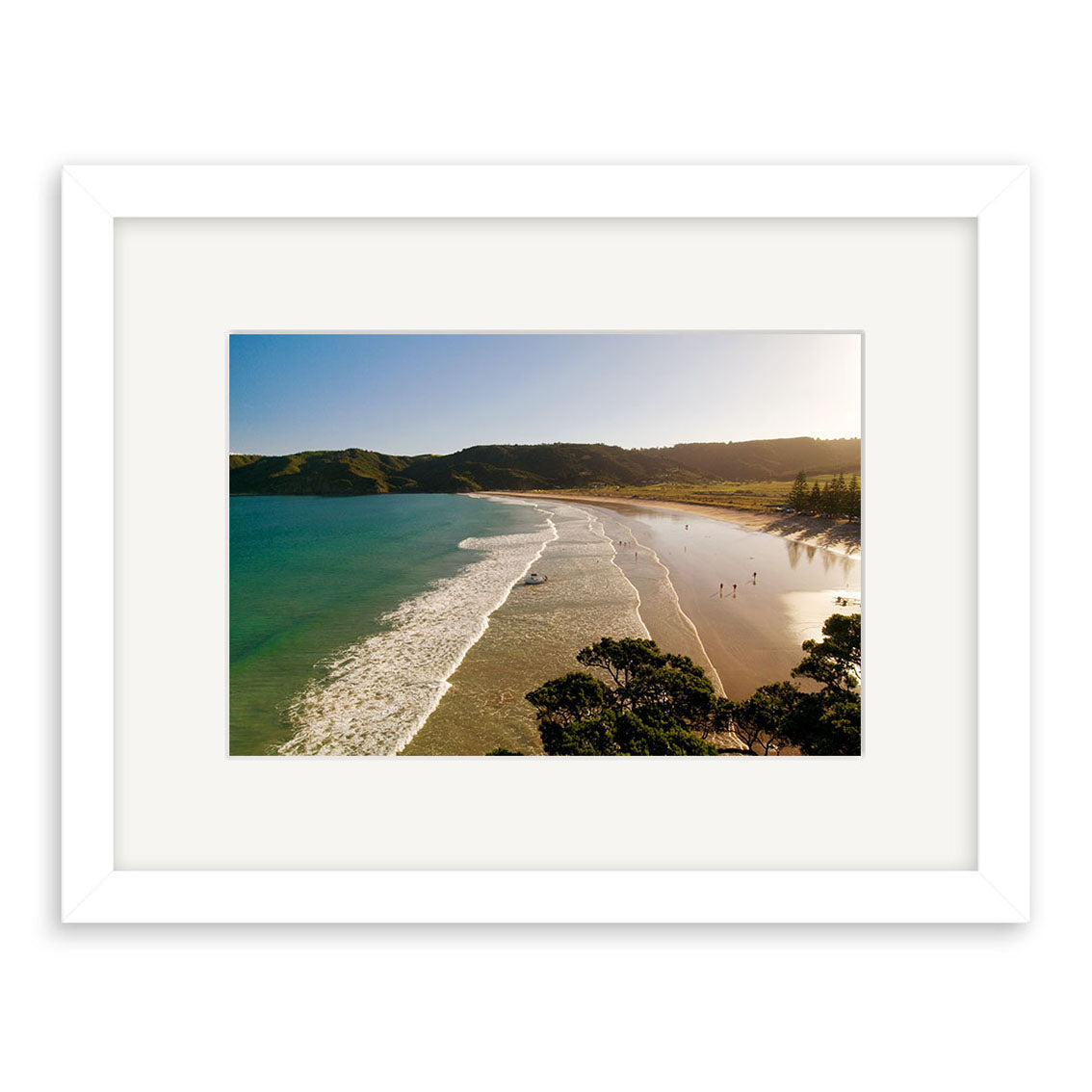 Framed photograph Matauri Bay - RP169 shows a sunny beach with gentle waves, people on the shore, and green hills. This Reuben Price print captures New Zealand scenery in a white mat and simple white frame.