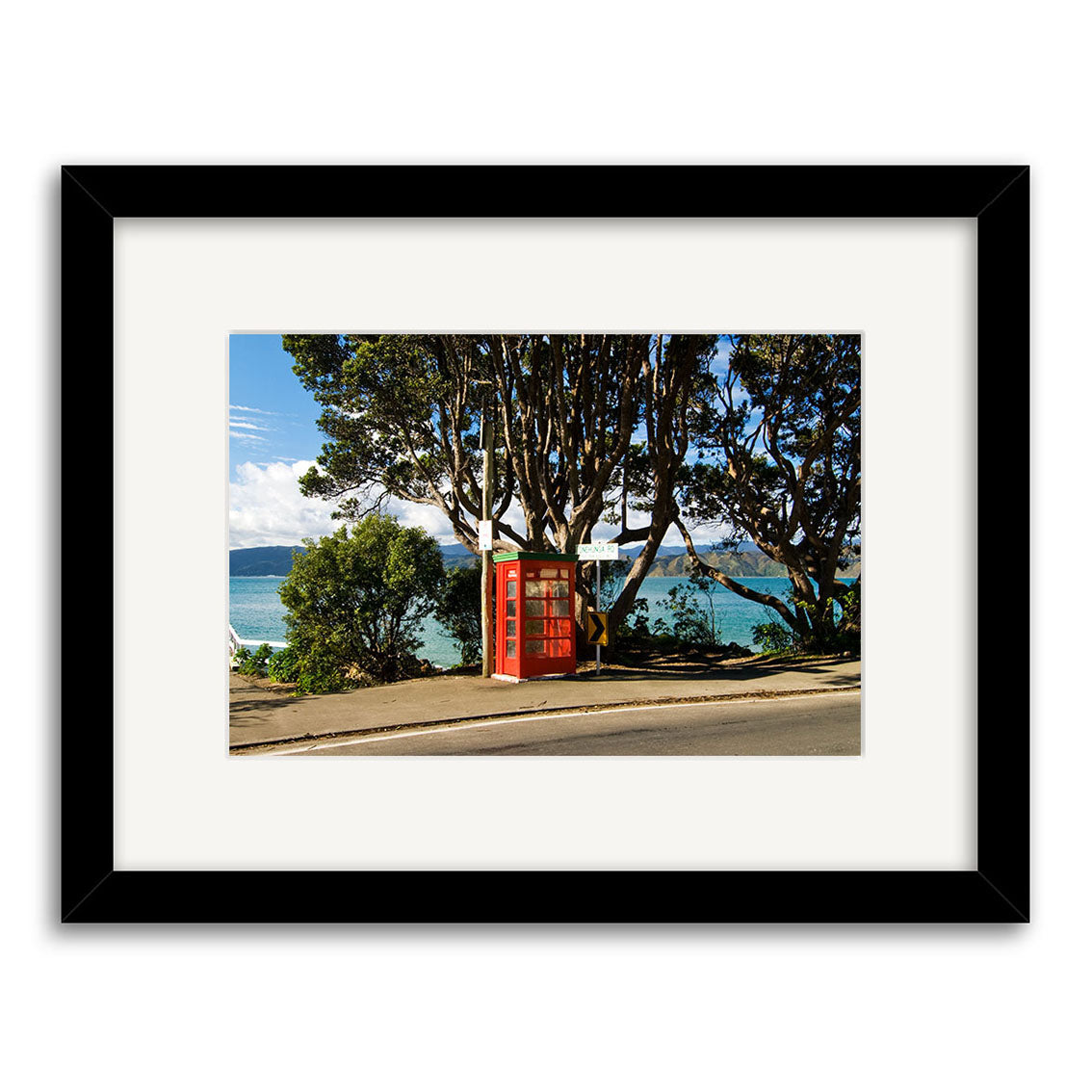 The Karaka Bay - RP170 photographic print features a classic red British-style phone booth on a New Zealand sidewalk near blue water and trees, set against a partly cloudy sky. It comes framed in black with a white mat.