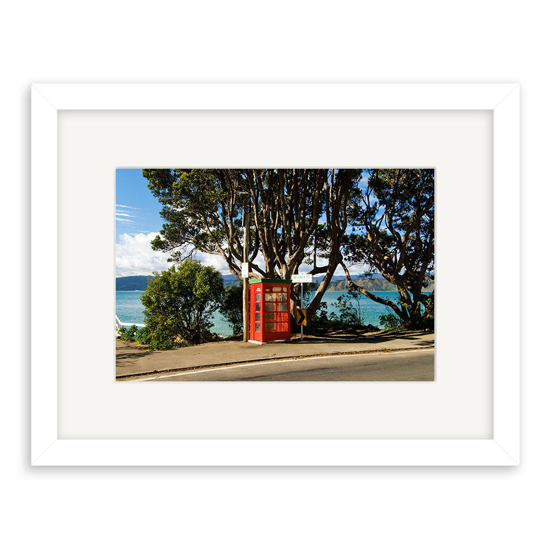 The Karaka Bay - RP170 print features a red British-style telephone booth by the roadside in New Zealand, with water, hills, and trees under a partly cloudy sky—beautifully captured on archival quality matte paper.