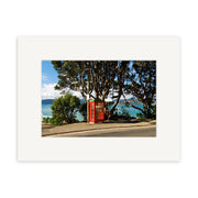 Karaka Bay - RP170 features a red phone booth on a sidewalk by the road, with trees, blue water, and New Zealand hills in the background under a partly cloudy sky—available as photographic prints on archival quality matte paper.