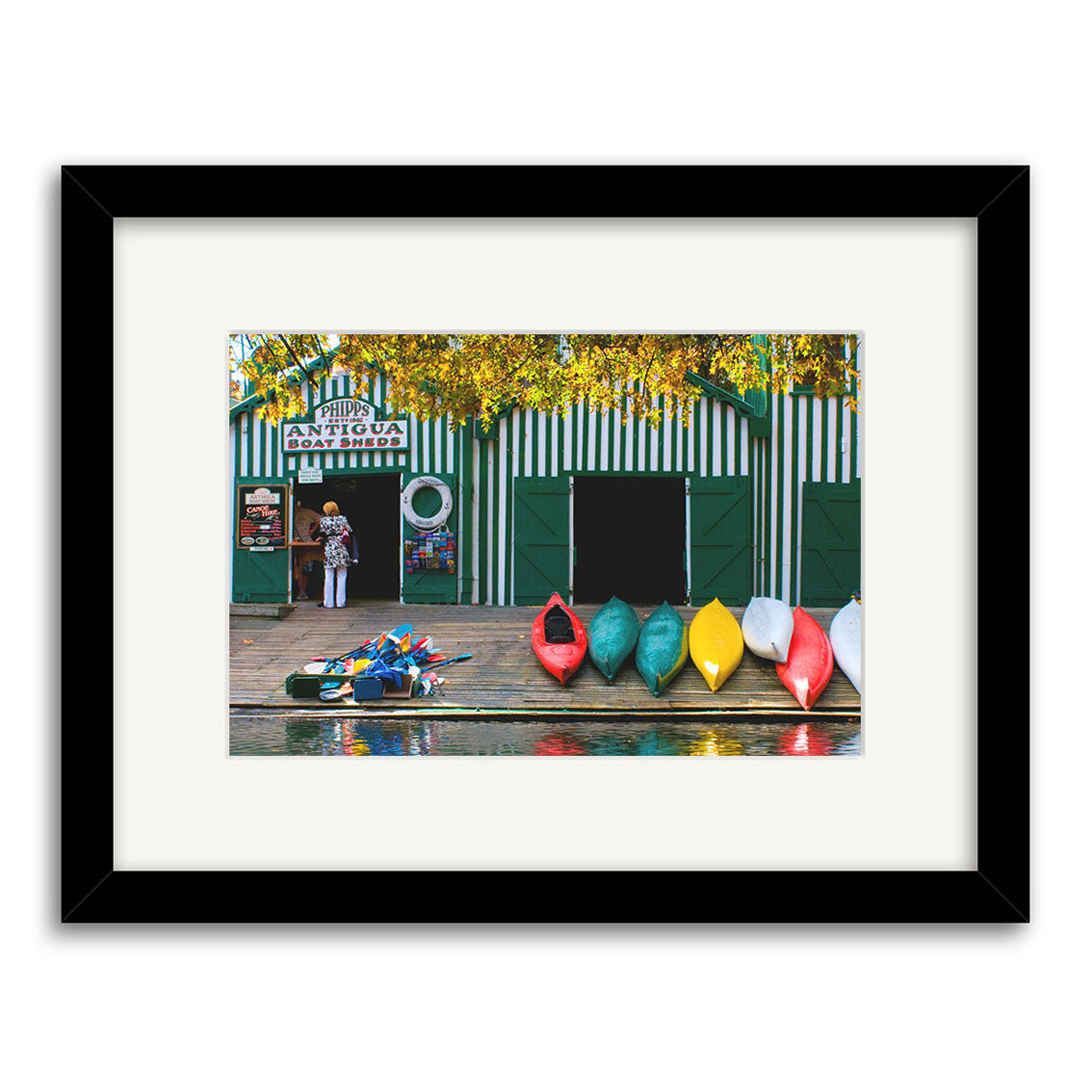 A green and white striped boat shed stands behind colorful kayaks by the dock, as a person stands under an Antigua Boat Sheds sign—featured in Reuben Price’s iconic photographic print, Antigua Boat Sheds - RP172.