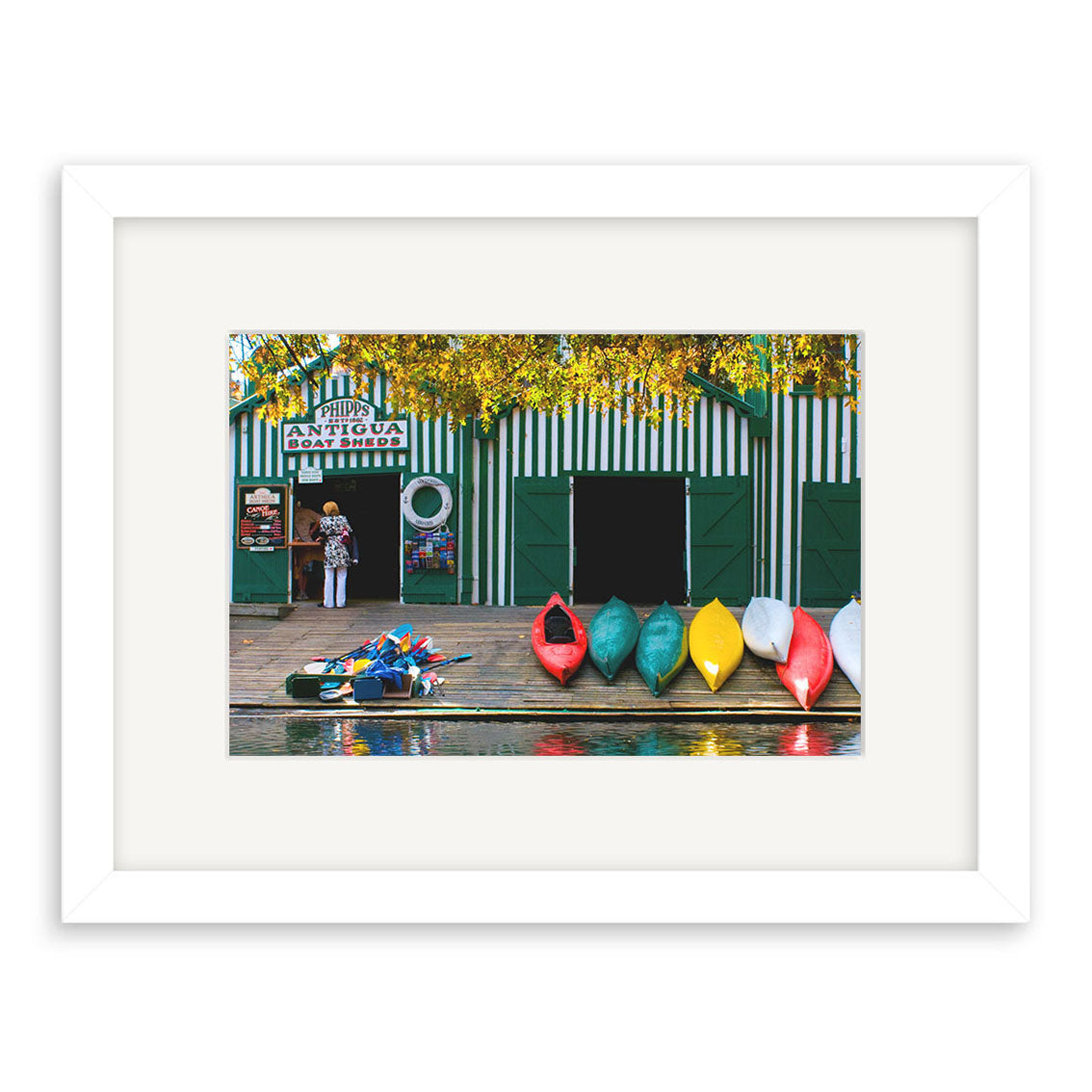 Antigua Boat Sheds - RP172: Framed print by Reuben Price, featuring a New Zealand boathouse with green-and-white stripes, a person at the entrance, and colorful canoes by the water beneath yellow-leaved trees.